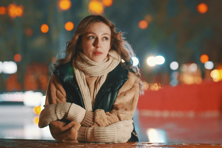 Blonde woman leaning on wall in scarf and gloves with wistful expression.