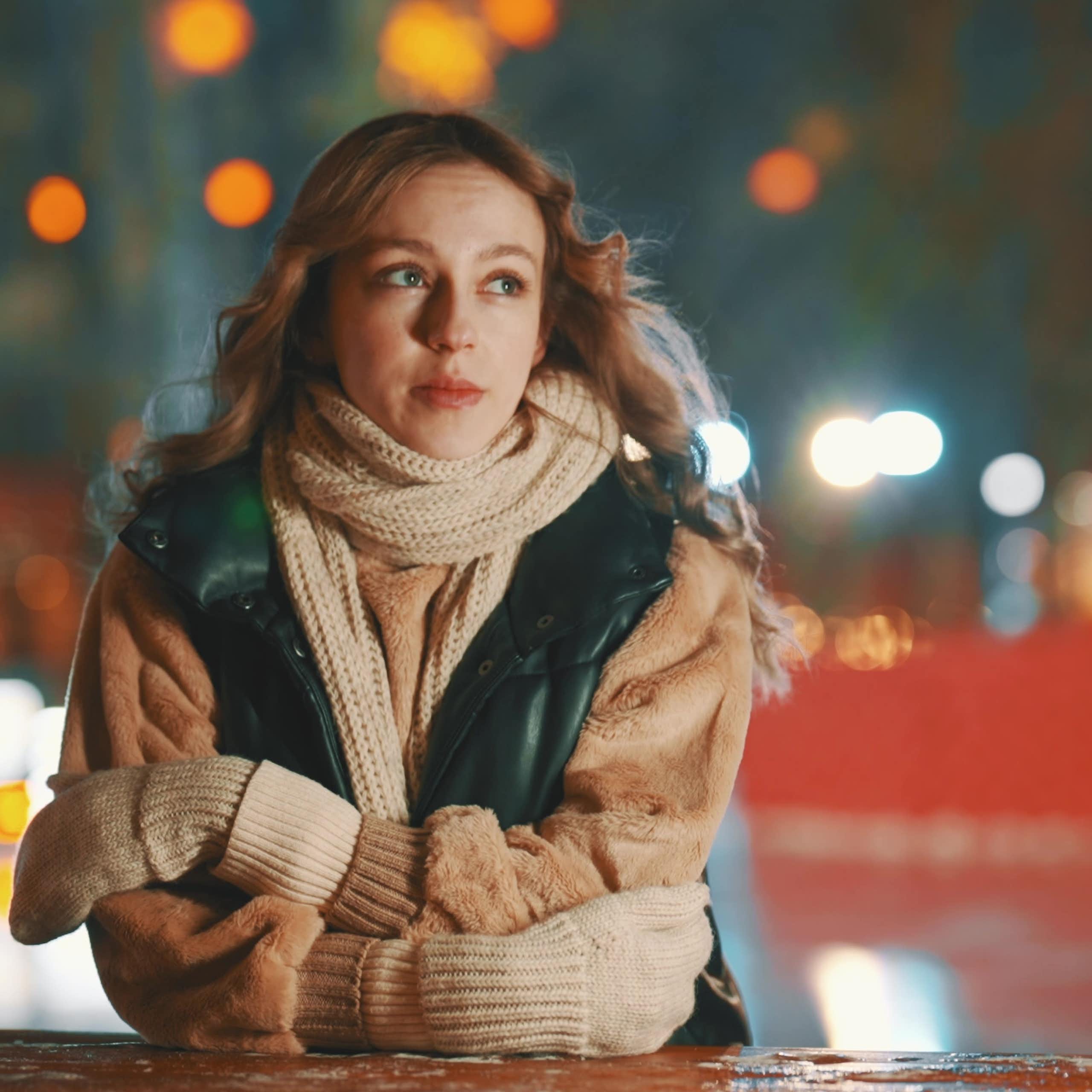 Blonde woman leaning on wall in scarf and gloves with wistful expression.