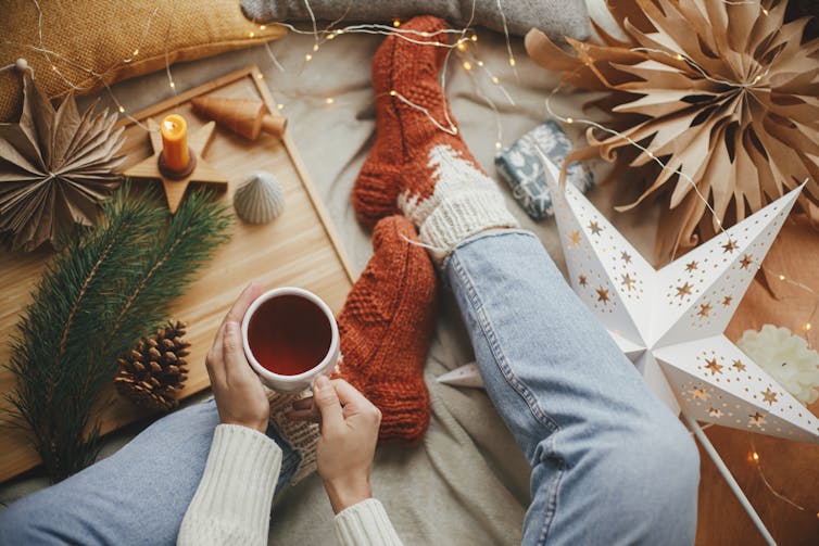 Close up of person sitting on floor with mug of tea surrounded by Christmas-y things.