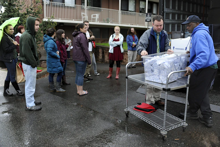 People stand around two men holding a bin of wrapped cheese.