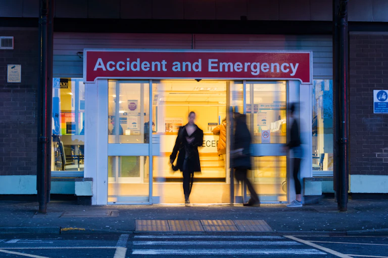The doors of an A&E at night. People are blurred to hide their identities.