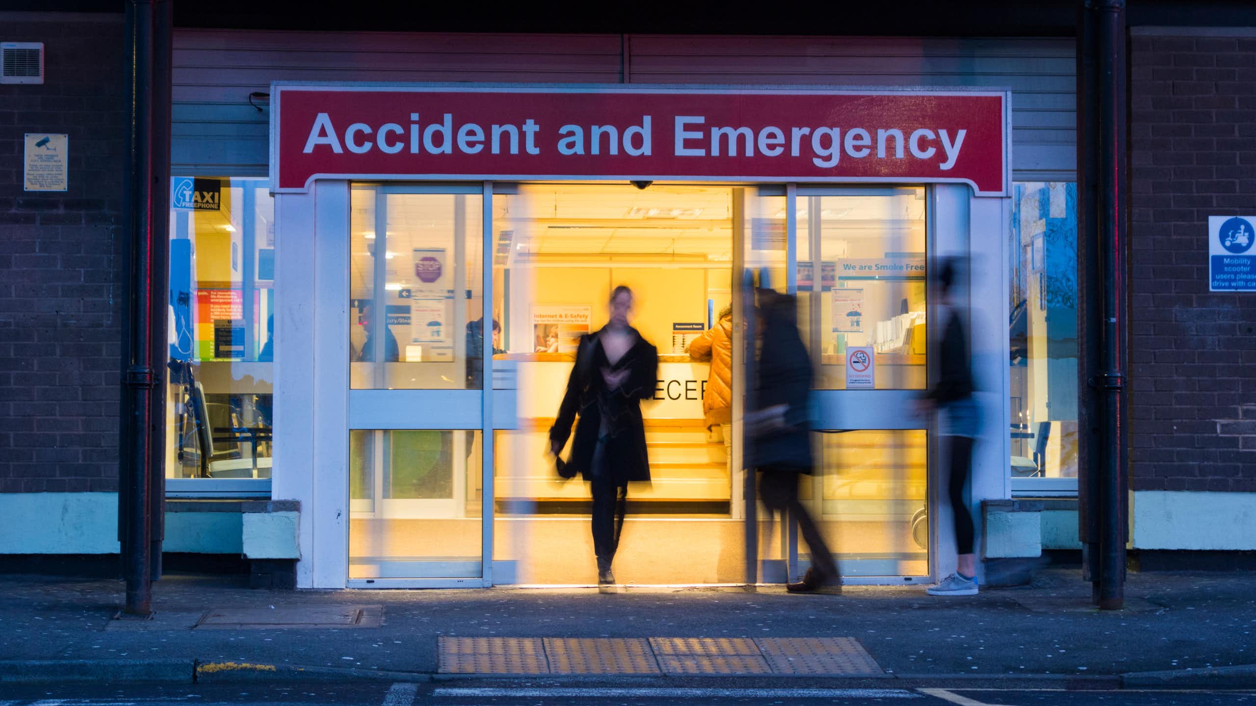 The doors of an A&E at night. People are blurred to hide their identities.