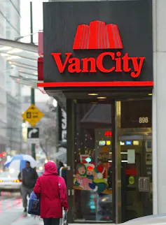 A person walks outdoors on a city sidewalk past a red sign that says Vancity.