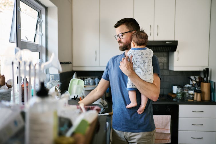 A brunette man with glasses holds an infant in one arm as he reaches into a sink in a cluttered kitchen.