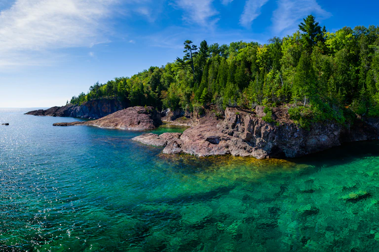 A view of the Lake Superior shoreline, with pine trees on sharp cliffs and wide expanse of blue water.