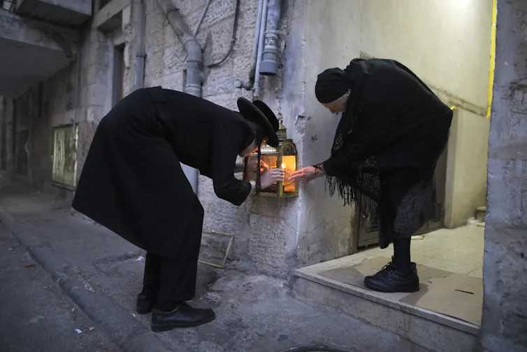 A man and a woman dressed in black bending down while lighting candles placed inside a glass box mounted on a wall.