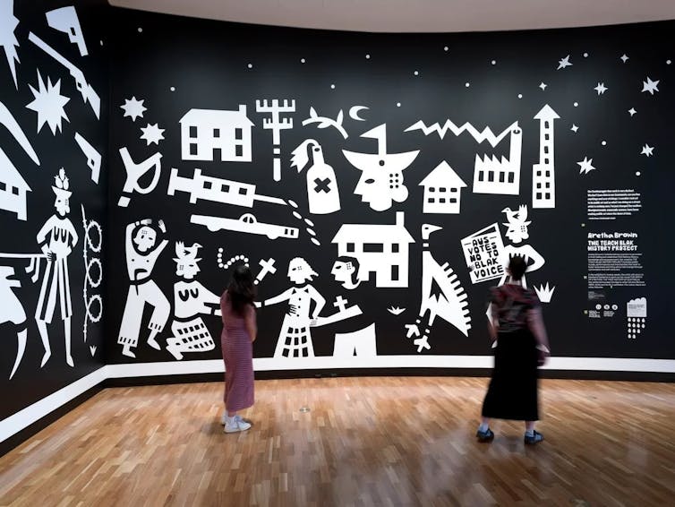 People stand in front of white paintings on a black background.