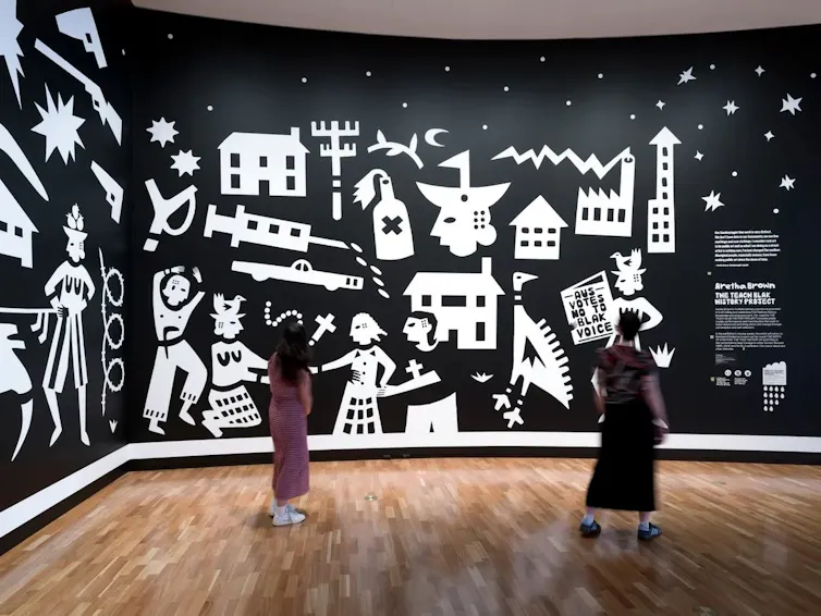 People stand in front of white paintings on a black background.