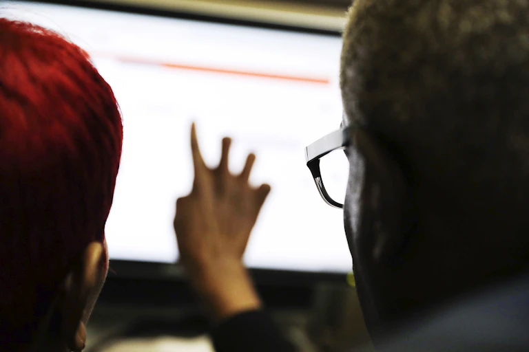 Two people are facing a bright computer screen. On the right is an African American man wearing glasses. On the left is a woman with red hair, and she is pointing at the screen.