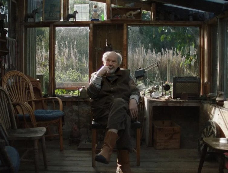 An older man sits on a chair in a dimly lit room.