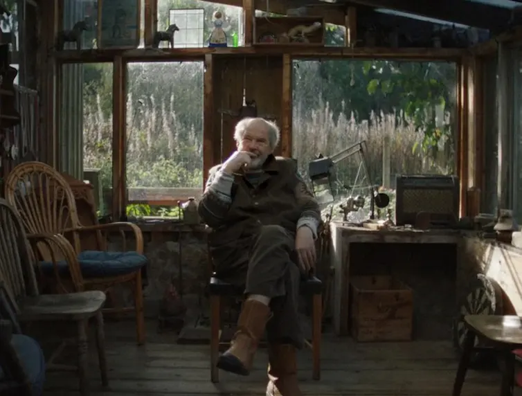 An older man sits on a chair in a dimly lit room.