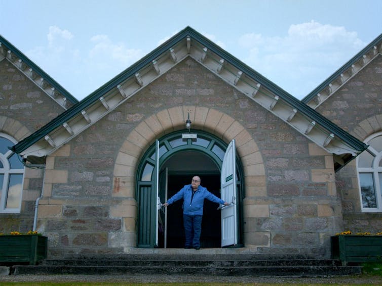 A man in a blue jacket stands in the open doorway of a stone building.