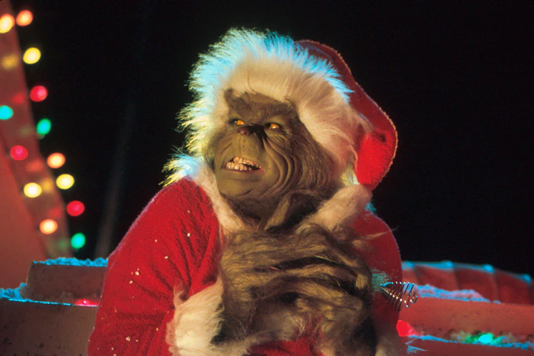 A movie still of Jim Carrey as The Grinch, wearing a red Santa hat and grinning diabolically.
