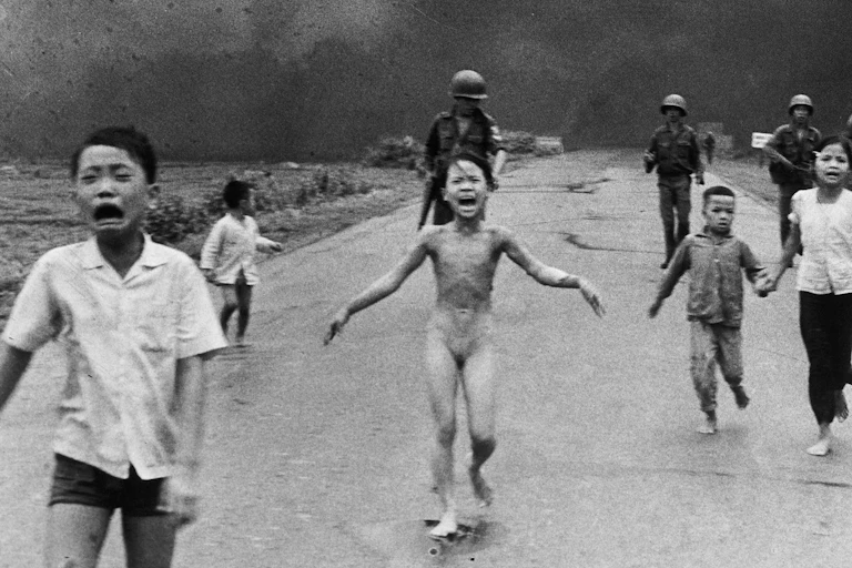 A black and white image shows a young girl, naked, running from a napalm attack during the Vietnam War, with other distressed children around her. Soldiers are visible in the background.