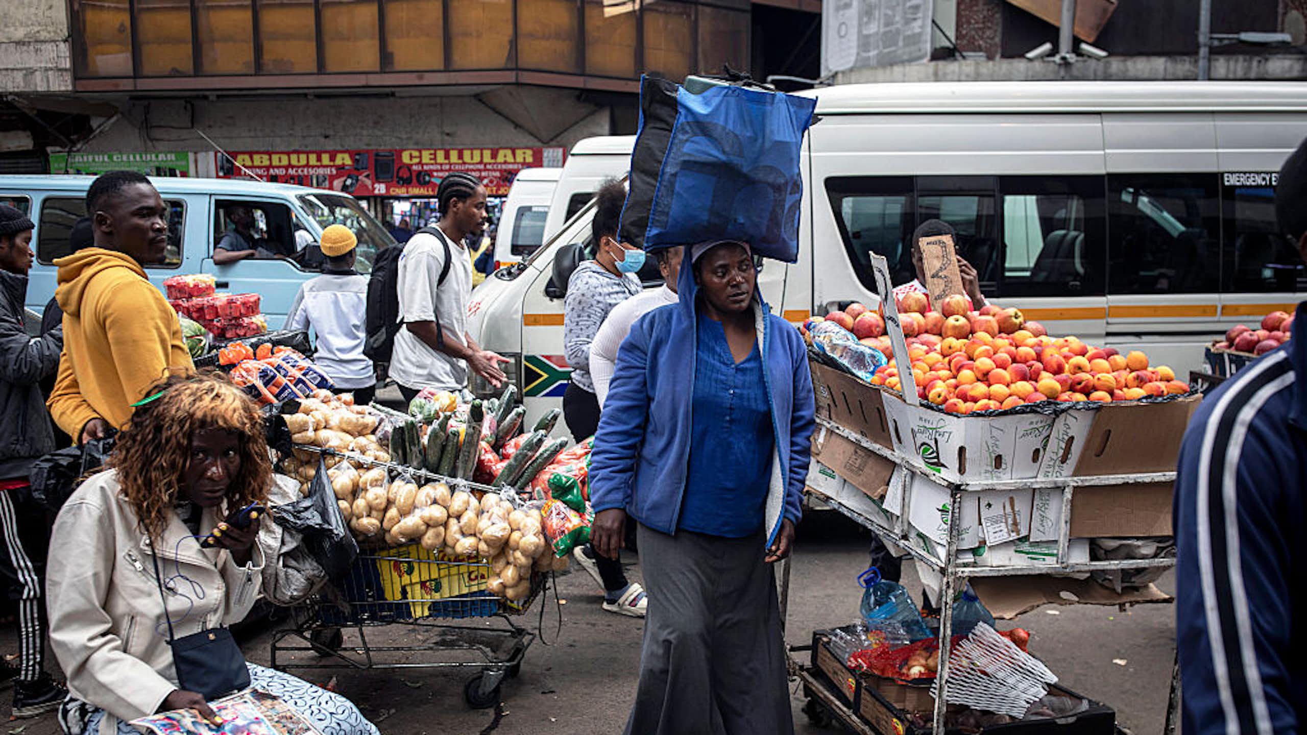 A woman walks with a package on her head past vendors selling fruit and vegetables in the street; minibus taxis in the background