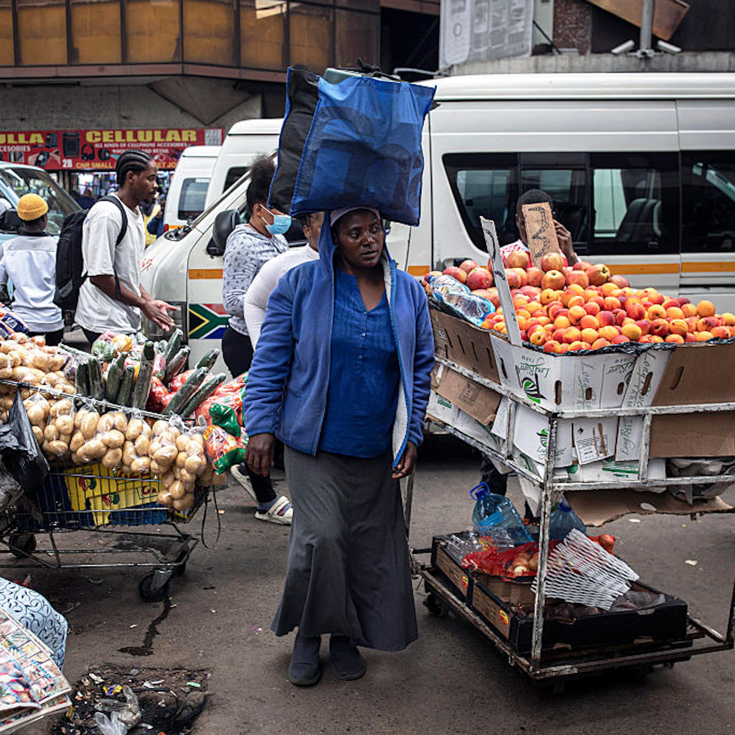 A woman walks with a package on her head past vendors selling fruit and vegetables in the street; minibus taxis in the background