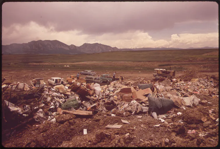 A broad swath of trash sits on the ground. In the distance are a green meadow and sharp mountain peaks.