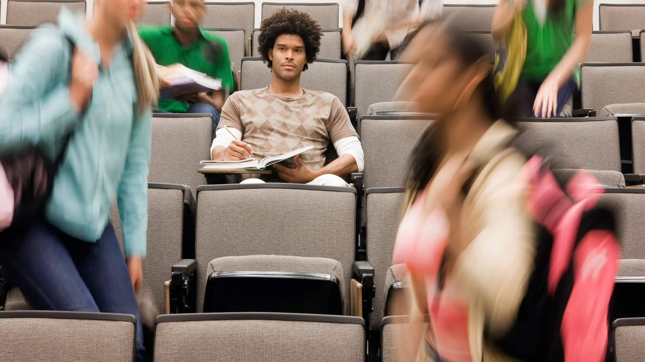 College student sitting in lecture hall while others walk around.