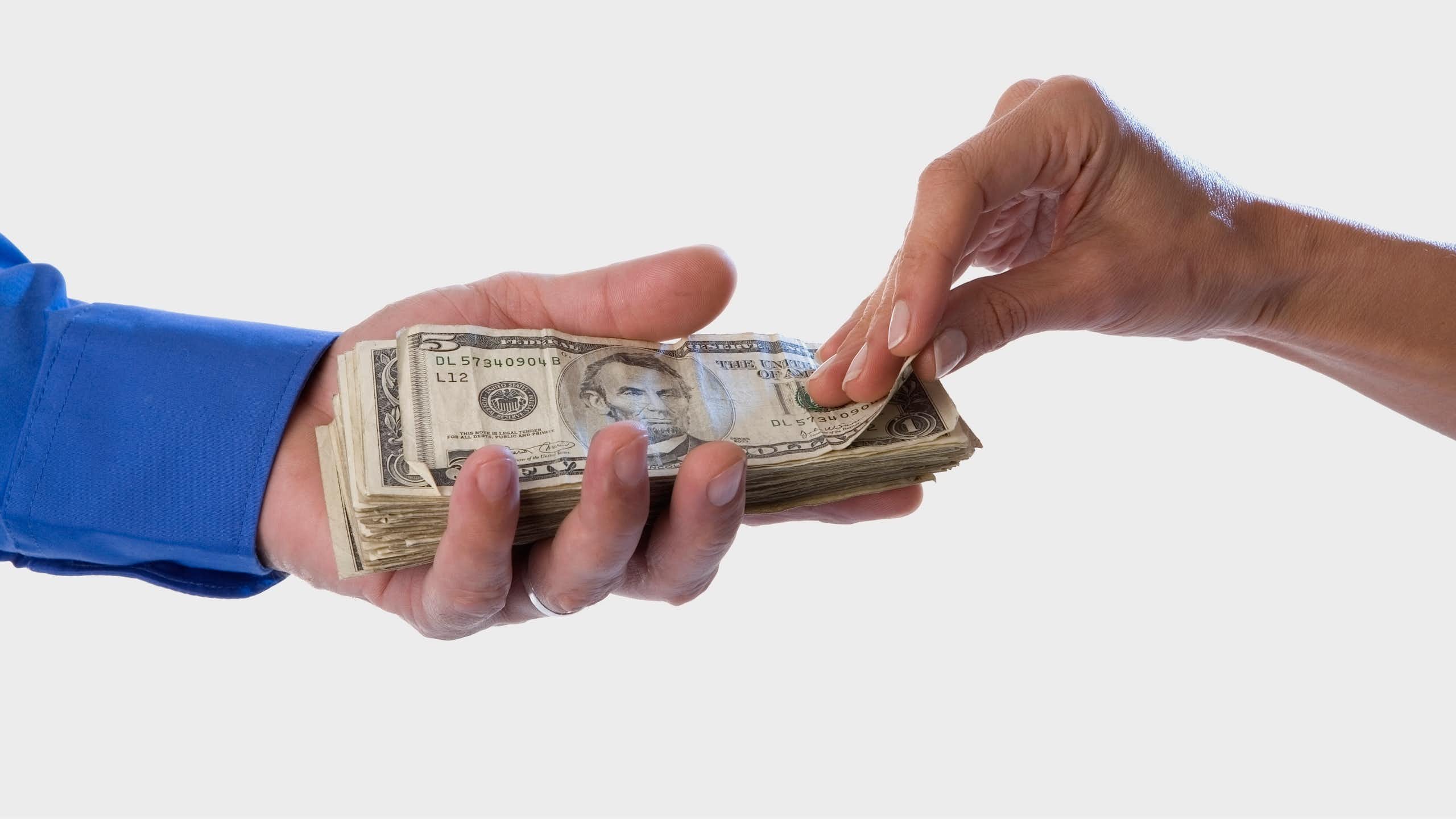 A hand with a blue sleeve holds a stack of USD cash as another hand pulls the top bill from the stack.