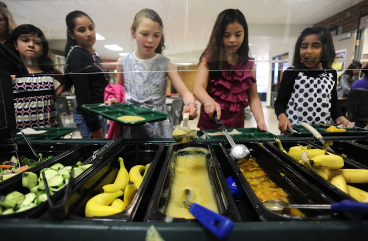 Children stand in front of a cafeteria line of food.