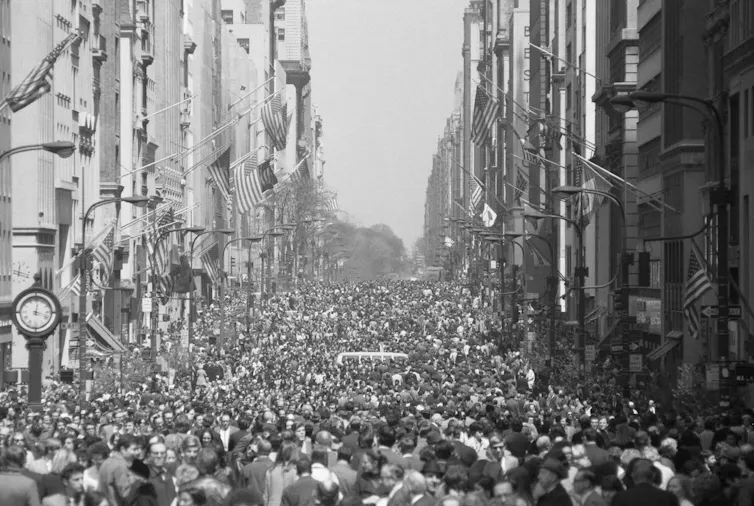Crowds of people cover all of a wide city street and its sidewalks.