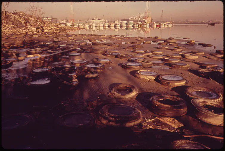 A beach covered in tires stretches out to a waterway, with docks and boats in the distance.