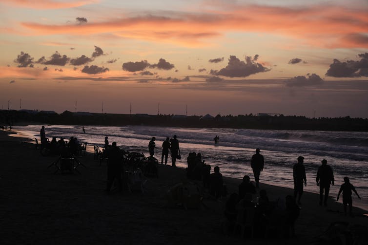 Pessoas vistas caminhando em uma praia contra um céu arroxeado.