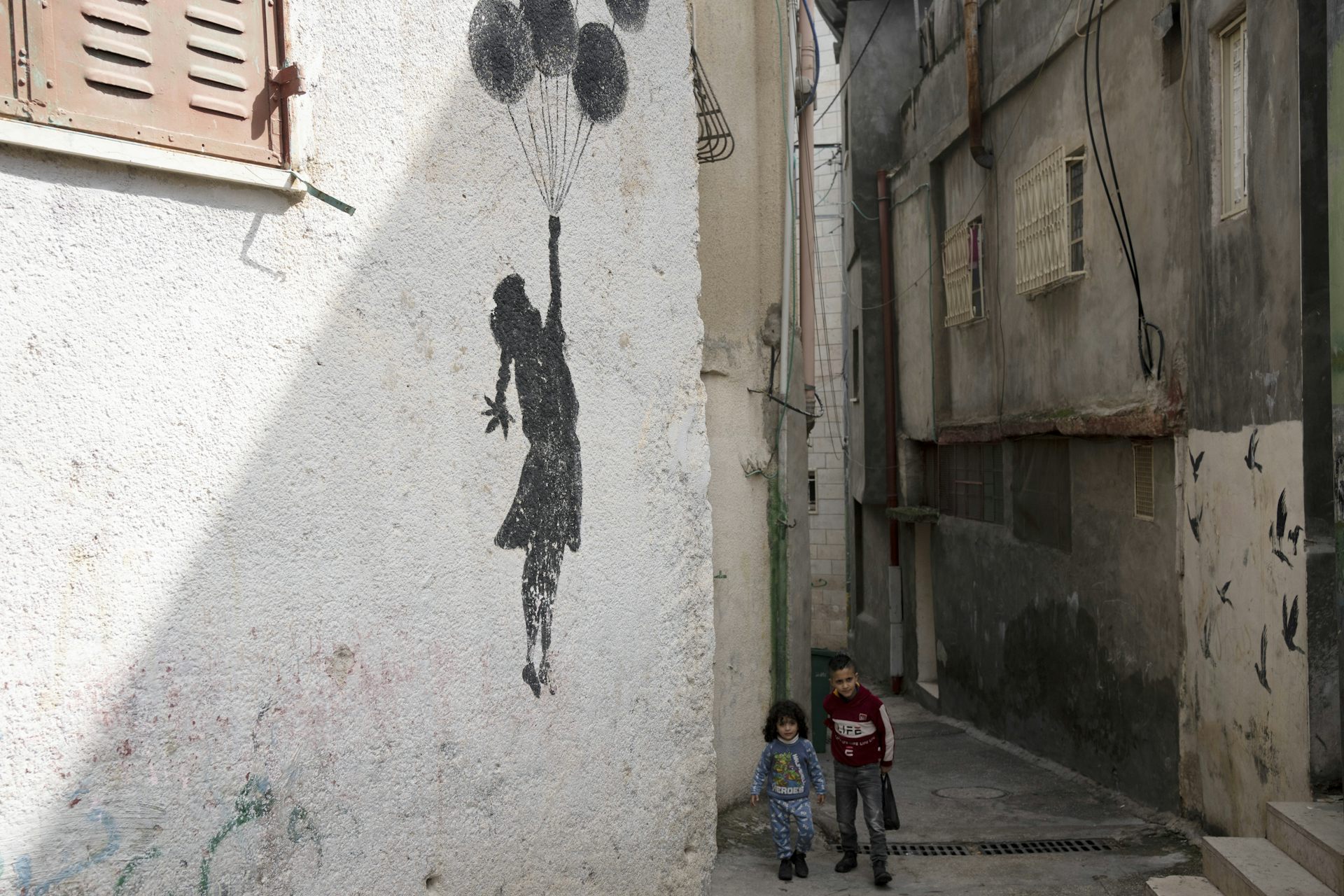 Two young children float up past a white wall with graffiti of a girl holding balloons on a string.