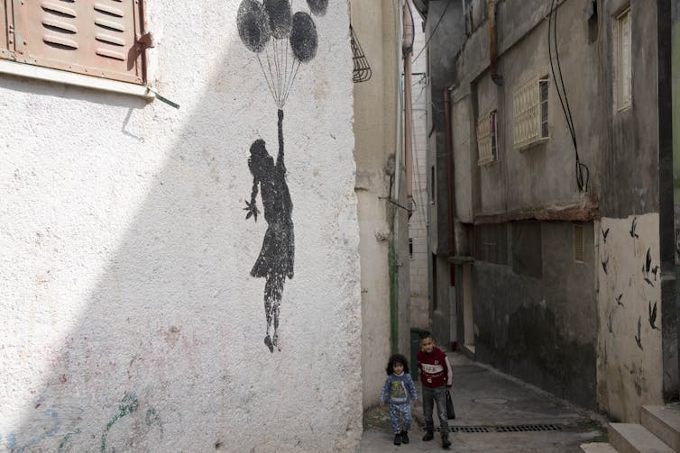 Two young children float up past a white wall with graffiti of a girl holding balloons on a string.
