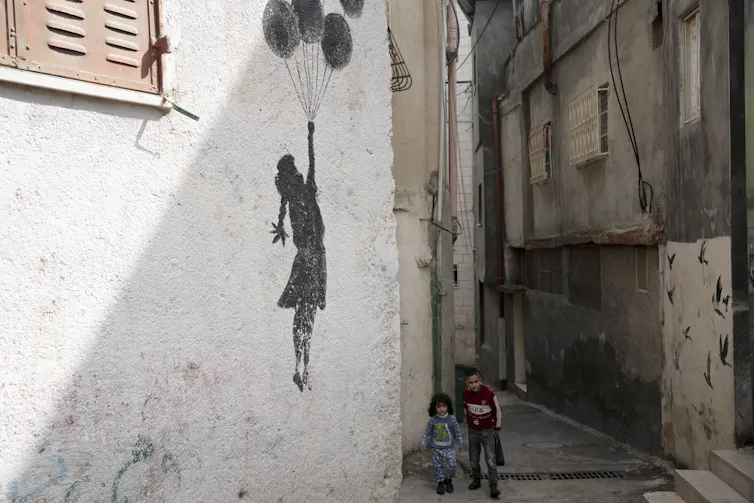 two young children walk passed a white wall with grafitti of a girl floating upwards holding balloons on a string
