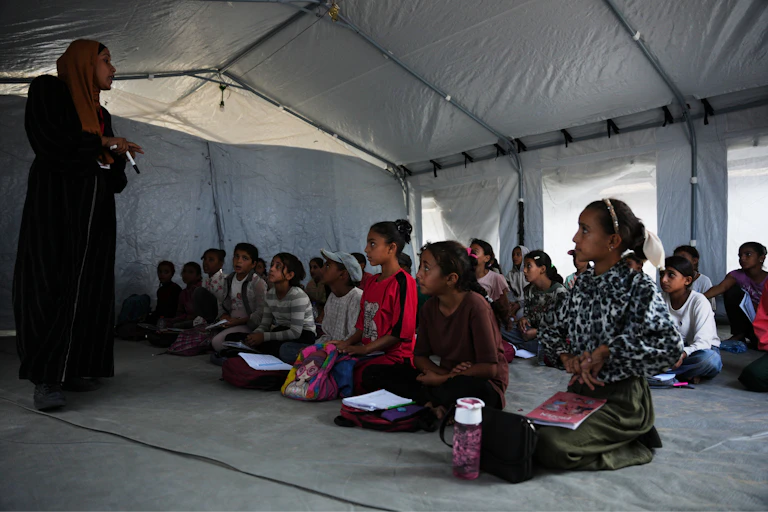 a woman wearing a hijab stands in front of a group of children seated on the ground inside a white tent.