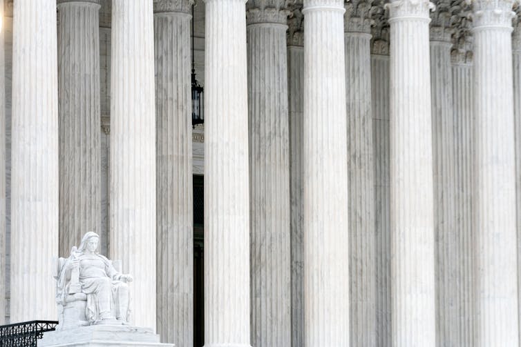 A statue of a woman, thinking, in front of the pillars of a large, white building.