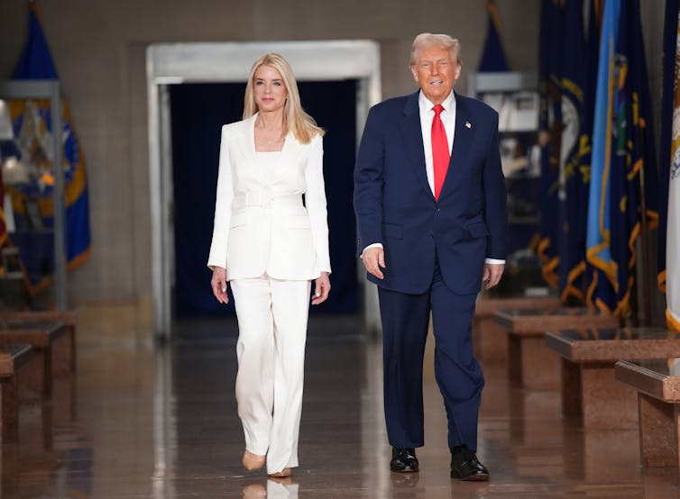 A woman in a white pantsuit walks next to a man in a blue suit, white shirt and red tie.