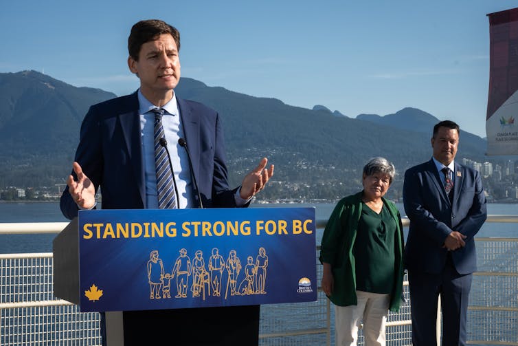 Un hombre con traje azul habla en un podio, una mujer con una chaqueta verde y otro hombre están parados detrás de él, detrás de ellos hay montañas en la distancia.