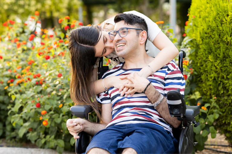 Woman has her arms around man sitting in a wheelchair and kisses him on the cheek