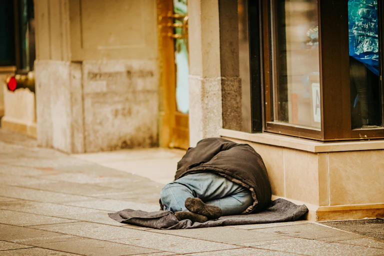 A homeless person is seen from behind, lying down on a grey blanket on a footpath.