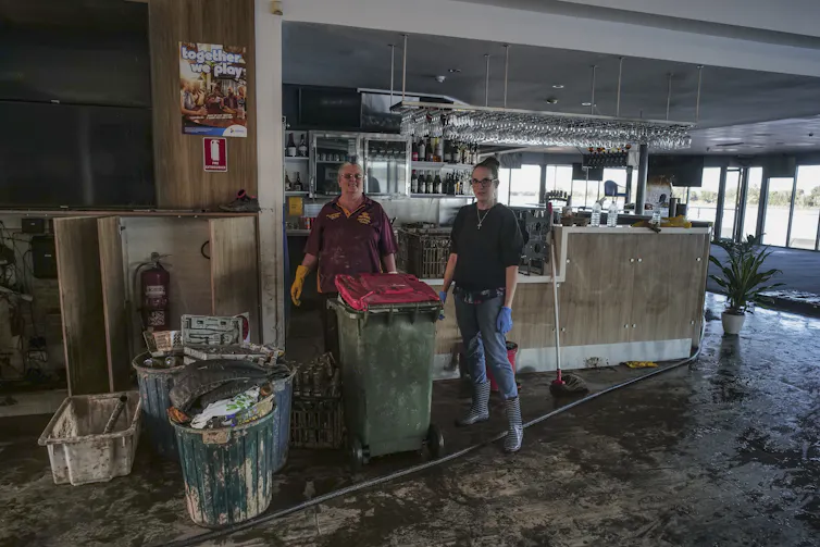 Two people stand in a building that has been recently flooded, showing mud on the floor.