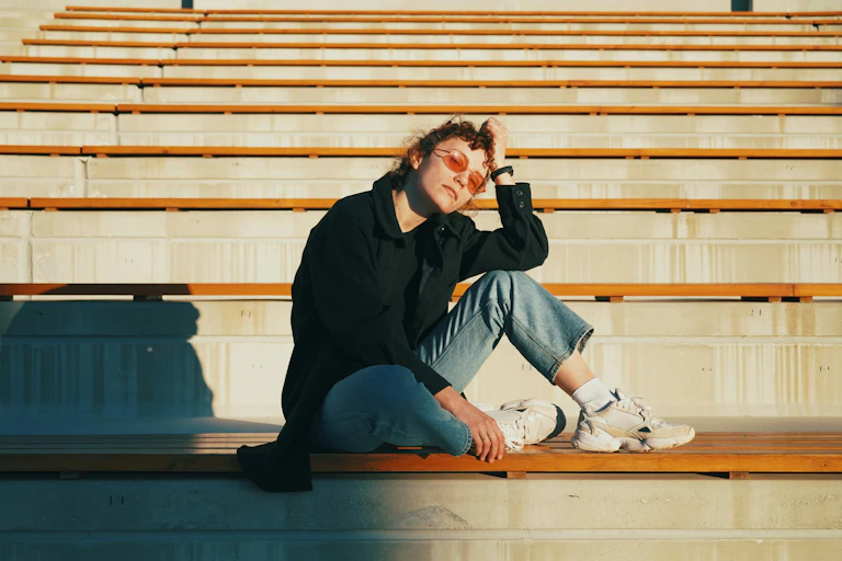 A young woman in sunglasses and chunky white sneakers sits on a bench