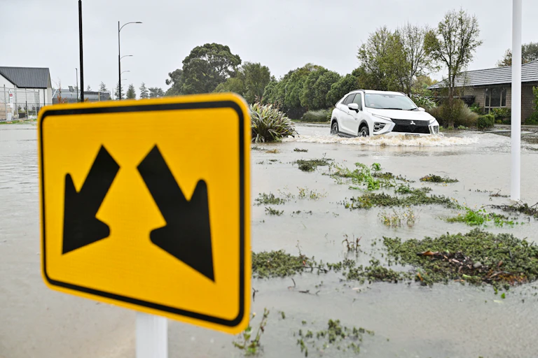 A flooding river, with a road sign in the foreground and a flooded car in the background.