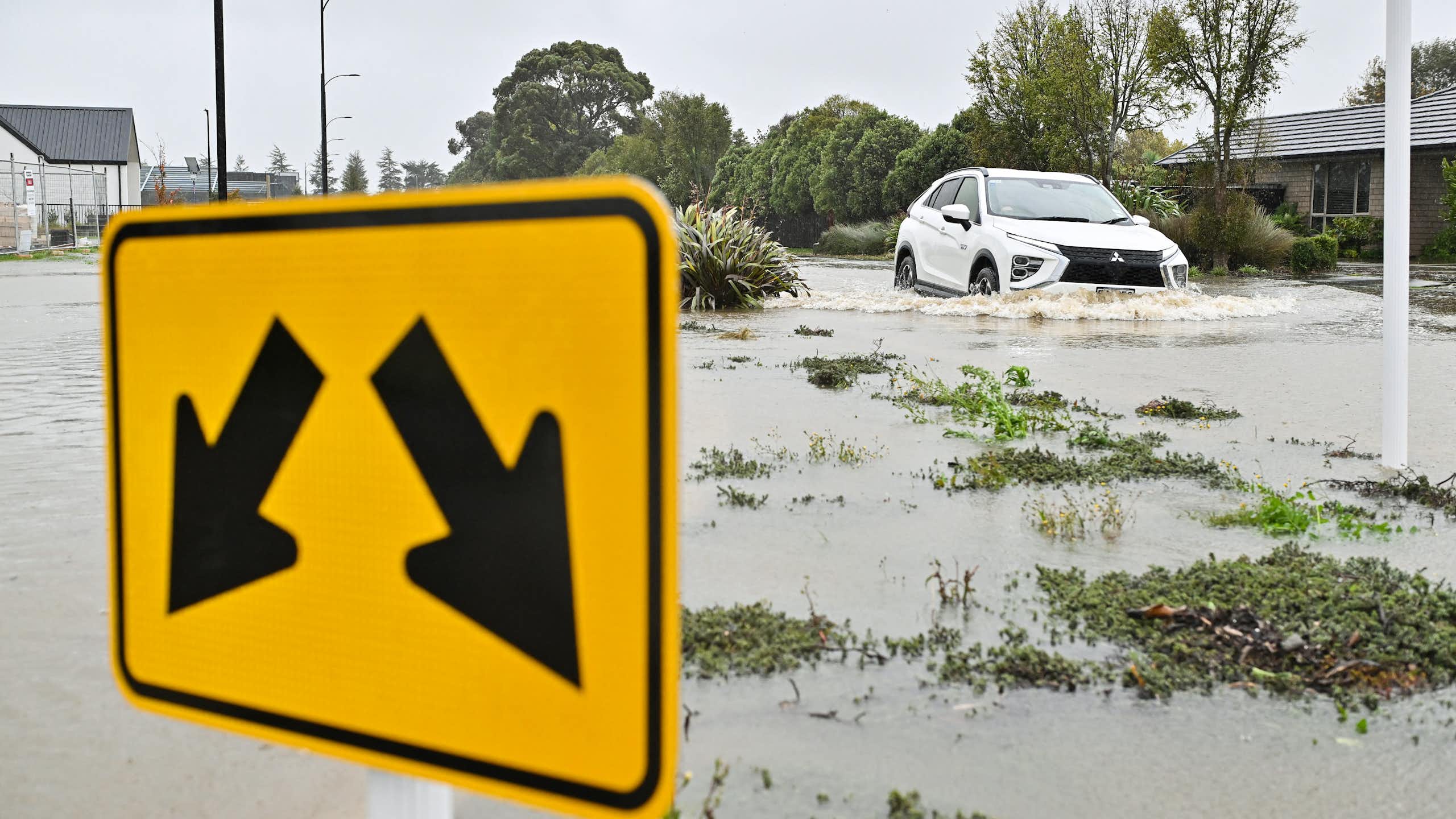 A flooding river, with a road sign in the foreground and a flooded car in the background.