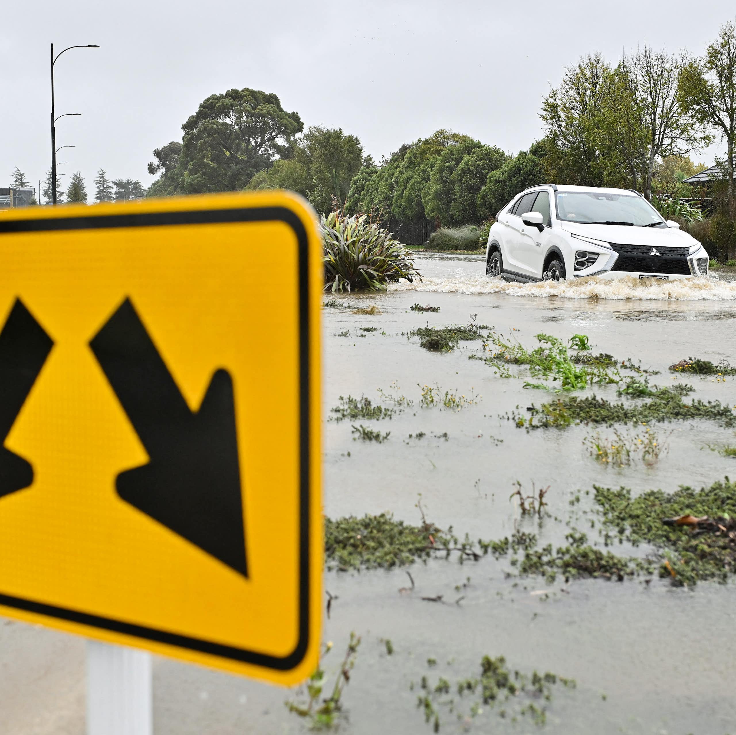 A flooding river, with a road sign in the foreground and a flooded car in the background.