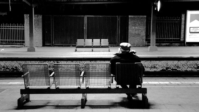 A man at a train station, sitting on a metal bench divided into four with armrests.