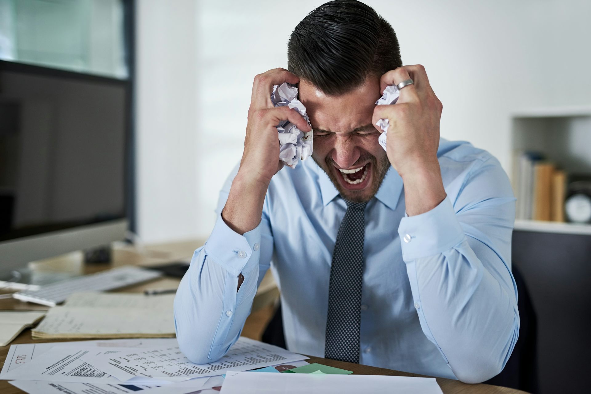 A man wearing a shirt and tie crumples two balls of paper and screams in frustration or anger while sitting next to his computer.