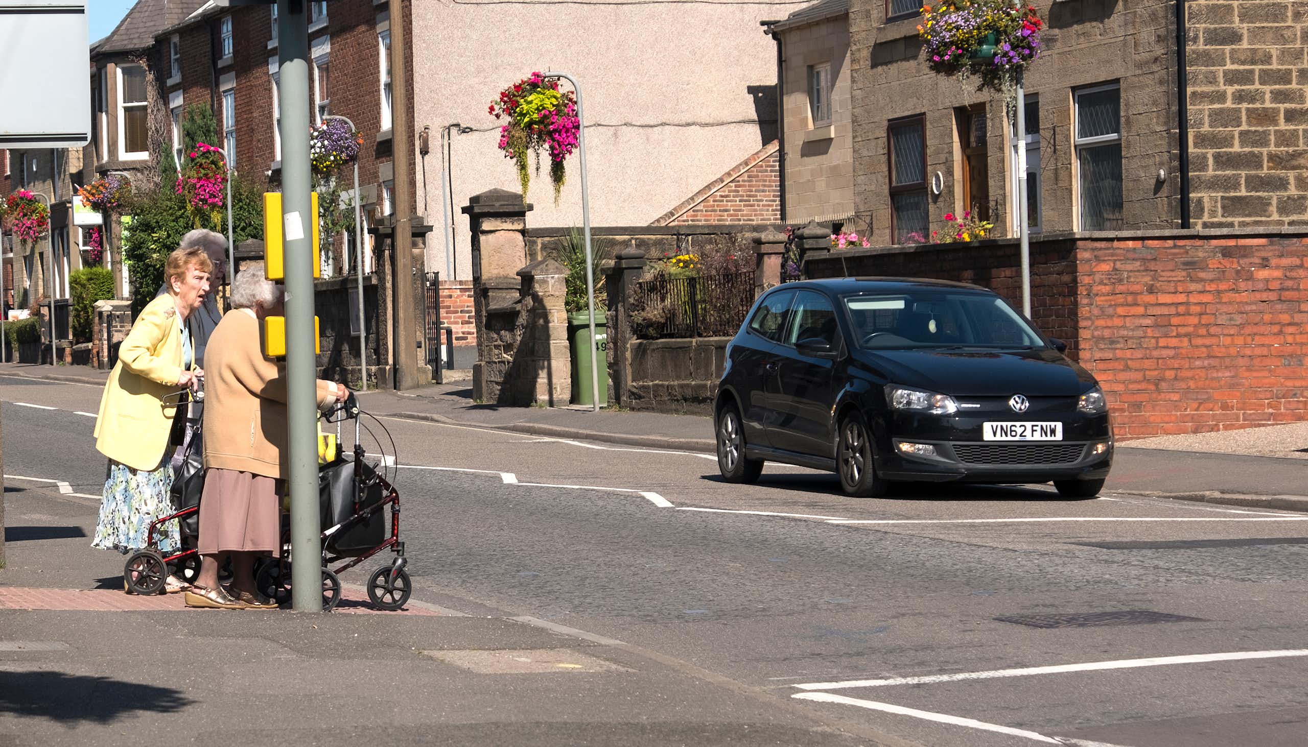 Three older people standing at a pedestrian crossing while a car waits.