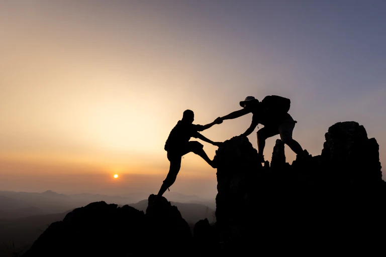 Silhouette of two people helping each other hiking up a mountain
