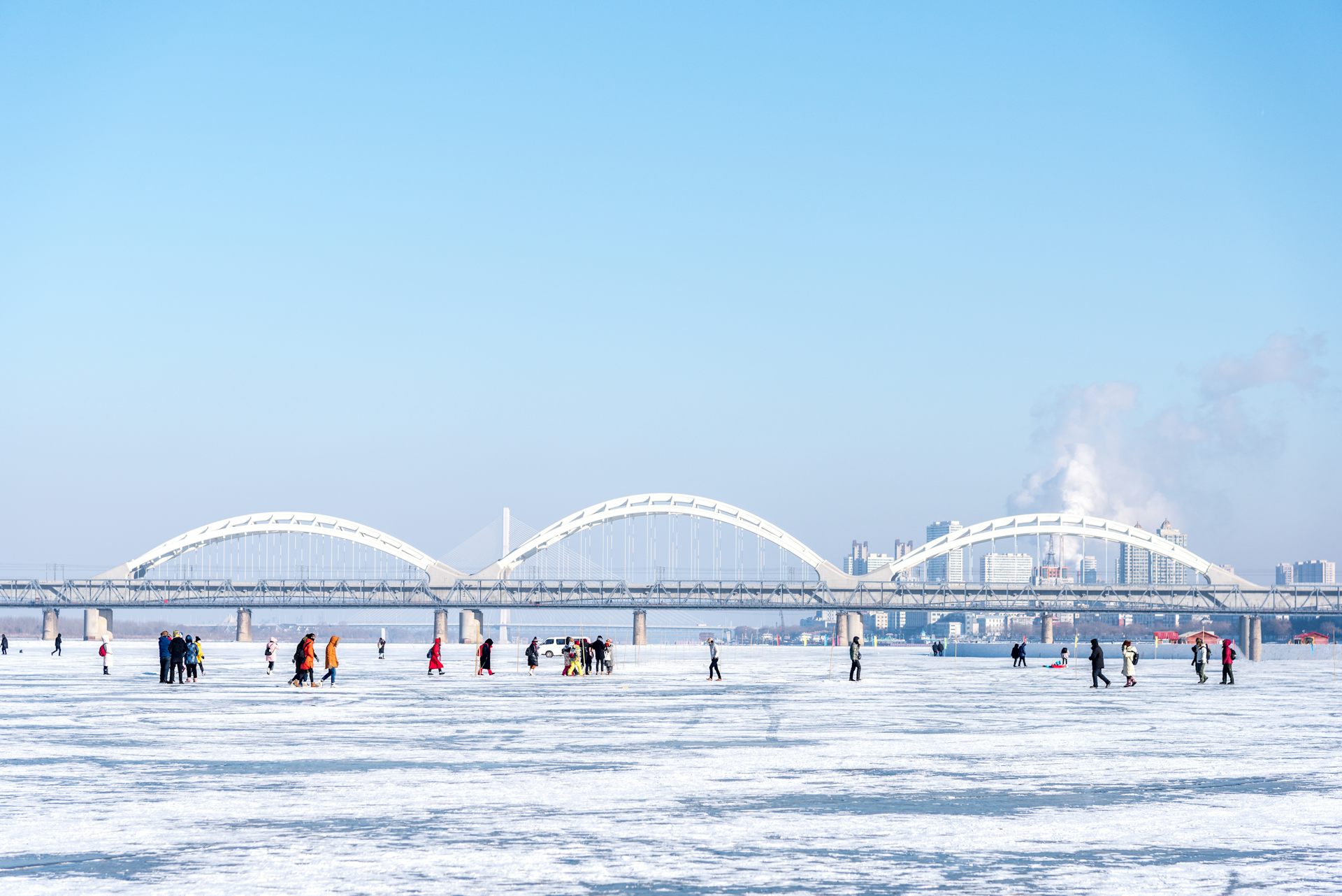 People walk on frozen river, bridge and city skyline in background