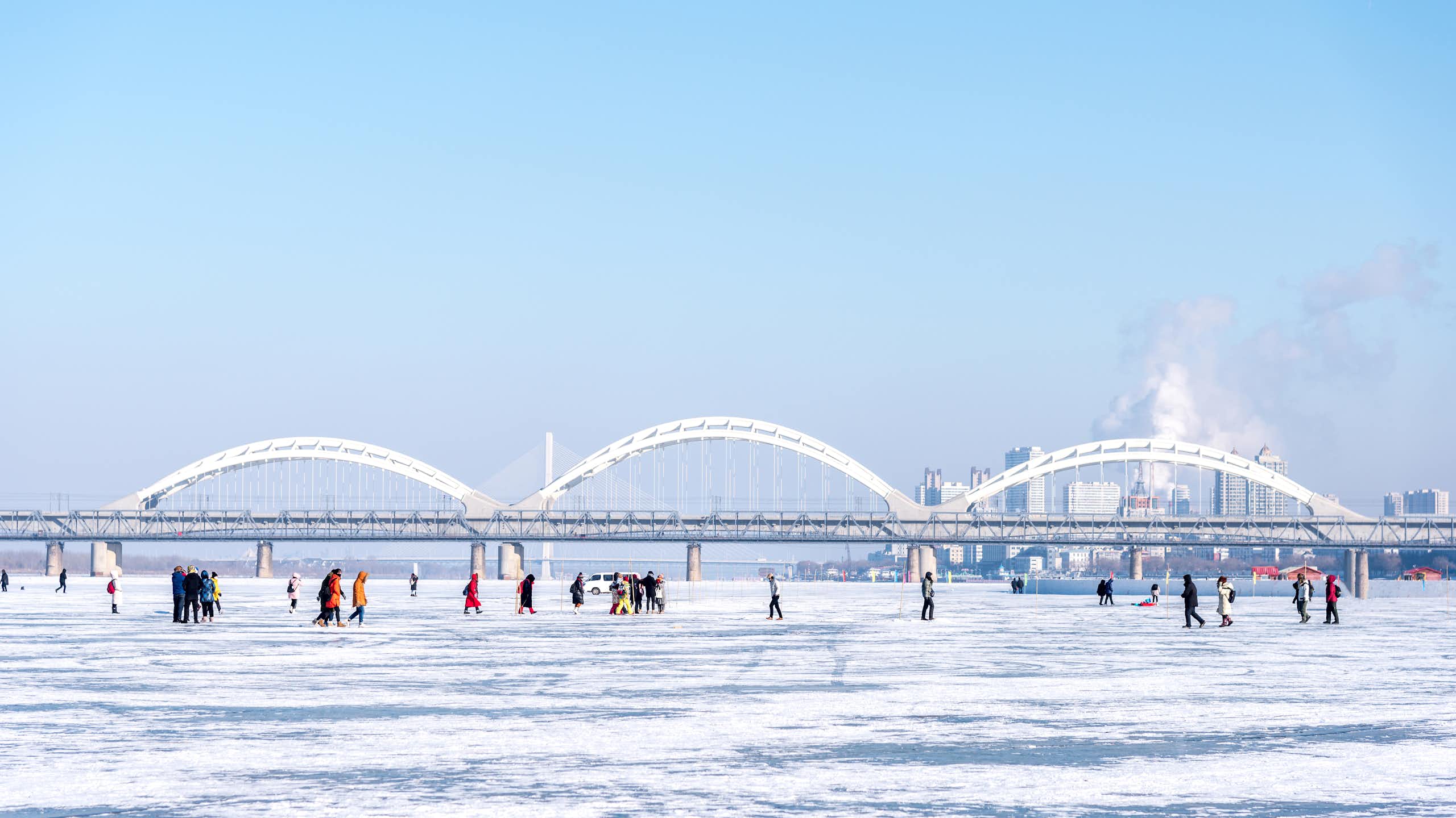 People walk on frozen river, bridge and city skyline in background