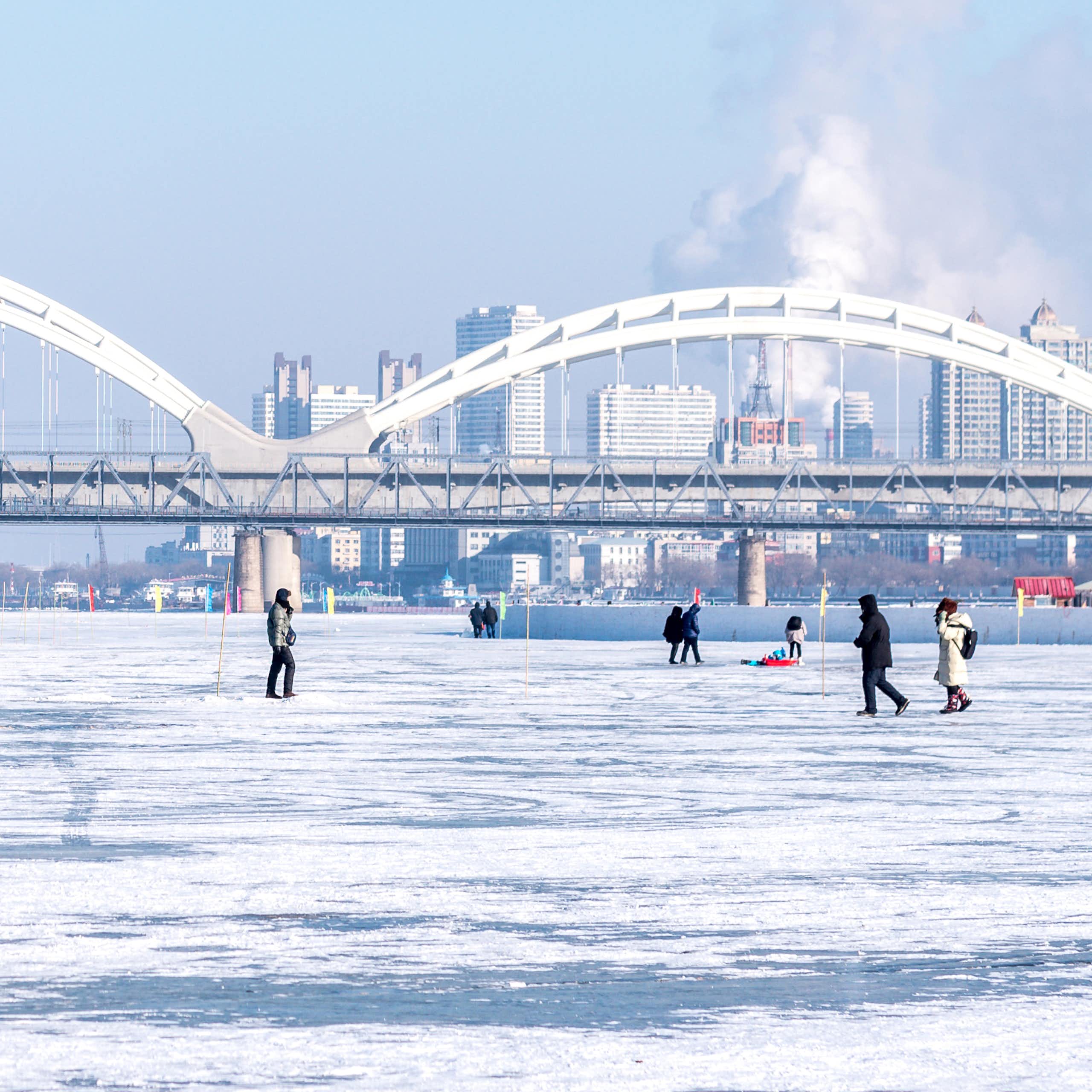 People walk on frozen river, bridge and city skyline in background