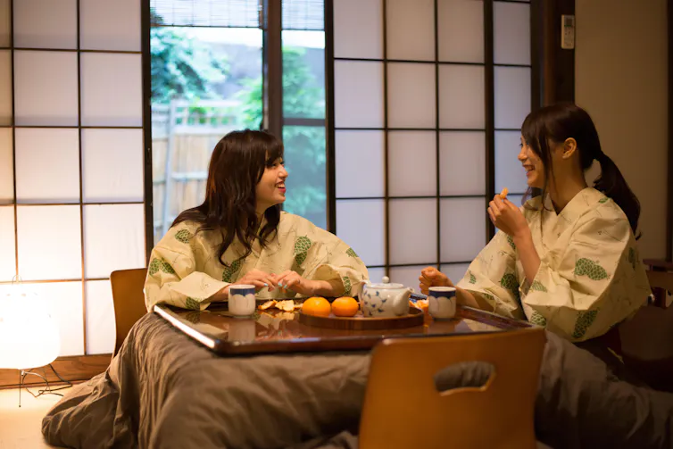 Two Japanese women sit at low table under blankets