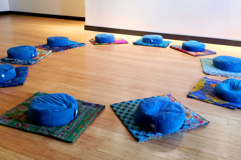 Several blue cushions are arranged in a circle on several mats laid out on a wooden floor.